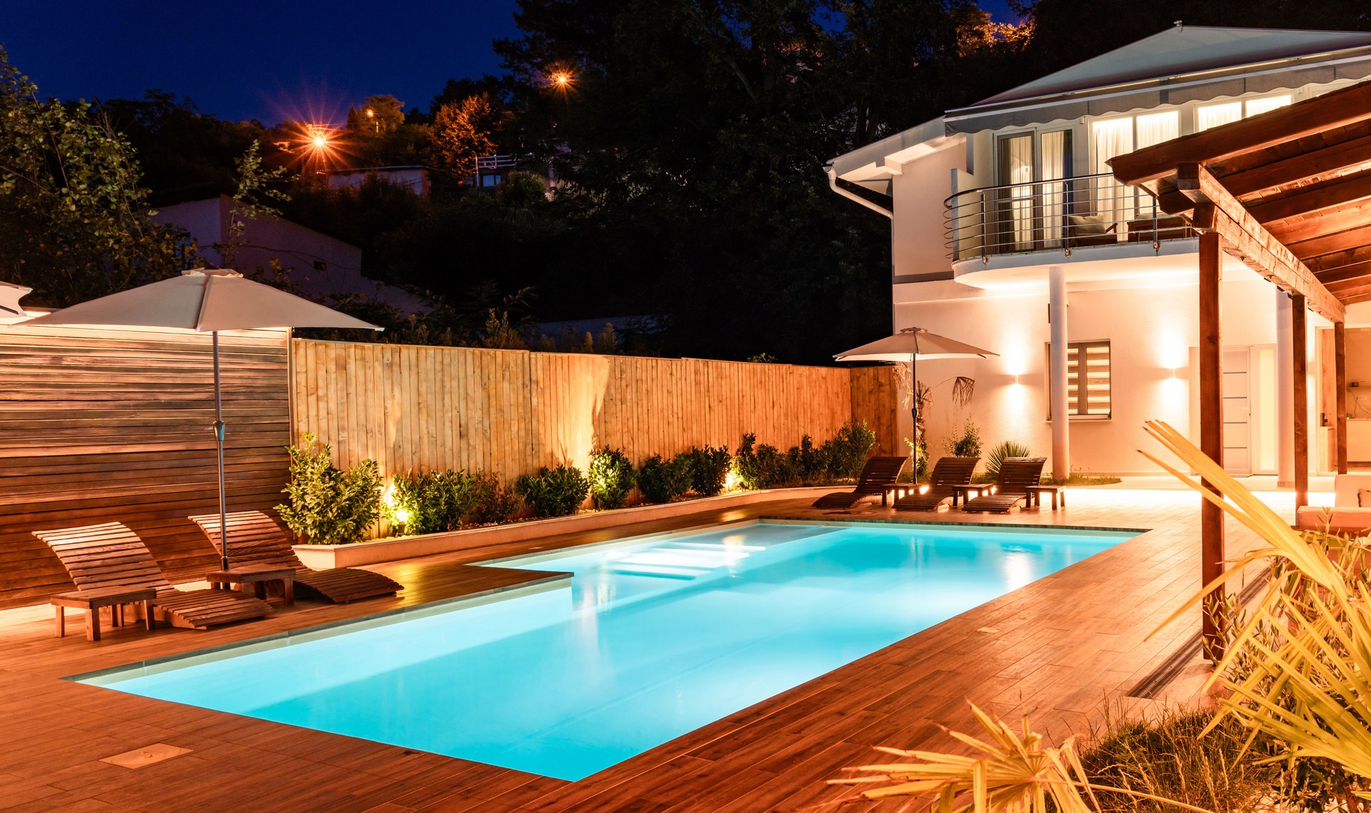 Elegant backyard with illuminated swimming pool and modern house at night. Luxury outdoor area with wooden deck and cozy lighting